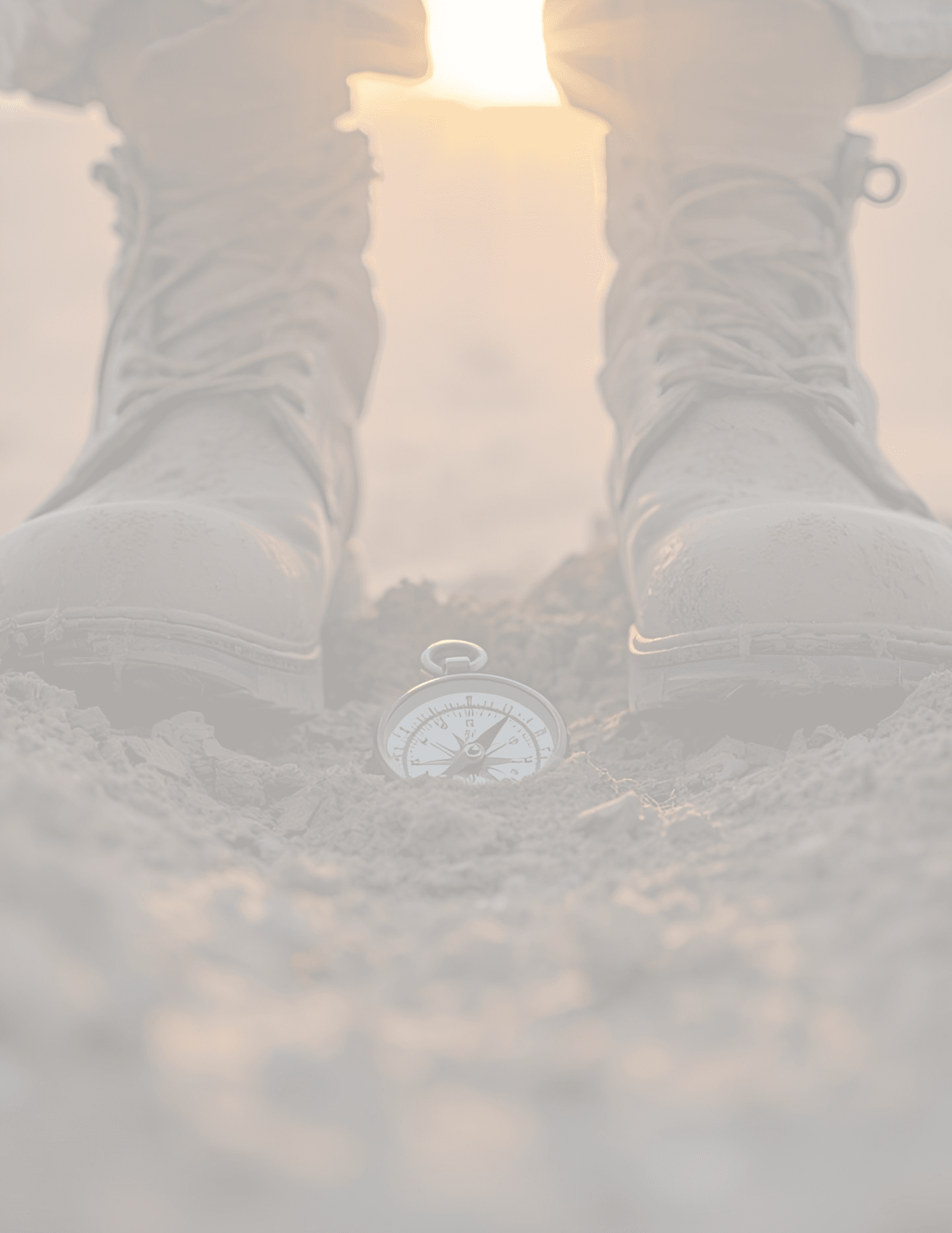A silver compass lies on rocky ground between two tan combat boots under bright sunlight.